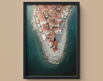 May include: Framed aerial photograph of a coastal town with red-tiled roofs and white buildings. The town is surrounded by turquoise water and a stone breakwater. The image is framed in a black border.
