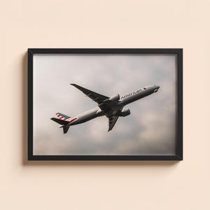 Op de afbeelding: Een ingelijste print met een vliegtuig in de lucht tegen een bewolkte lucht. Het vliegtuig is zilverkleurig met het woord "American" op de zijkant en rood-wit-blauwe staartvinnen. De print zit in een zwart frame.