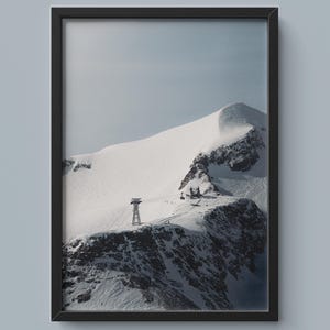 Può includere: Stampa incorniciata di una montagna innevata con cielo azzurro chiaro. Una funivia e degli edifici sono visibili sulla cresta. La cornice è nera.