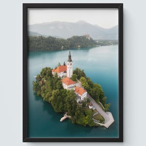 May include: Framed print of Lake Bled, Slovenia, with a church on an island. The image depicts a scenic landscape with a church, red-tiled buildings, and green trees surrounded by turquoise water and mountains in the background.