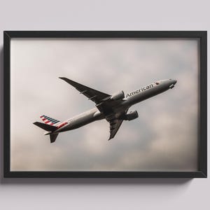 May include: A framed print featuring an airplane in flight against a cloudy sky. The aircraft is silver with the word "American" on the side and the American flag design on the tail. The print is in a black frame.
