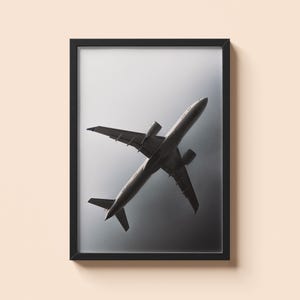 May include: Framed monochrome print of an aeroplane viewed from below. The aircraft is centrally positioned, with wings and tail visible against a cloudy sky. The image is in a black frame.