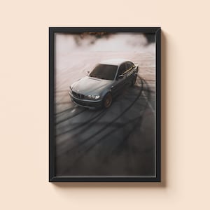 May include: A framed print showing a grey saloon car drifting on a track, with tyre marks visible. The car is central, with a blurred background of smoke and asphalt. The print is in a black frame, mounted on a beige wall.