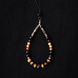 Op de afbeelding: Een kralen telefoonhanger met een lus van tijgeroog, zwarte en lichtbruine houten kralen, geaccentueerd met kleine zilverkleurige tussenstukjes. De hanger heeft een zwart koordje en is onderaan voorzien van de tekst 5610 The Pop-up.