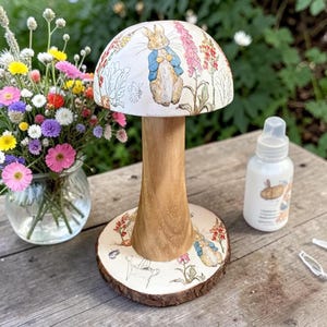 May include: A decorative wooden mushroom-shaped stand featuring a white cap and base adorned with illustrations of Peter Rabbit and colourful flowers. The central stem is made of natural light brown wood with a visible grain, standing on a round base.