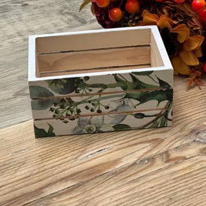 May include: A small, rectangular wooden crate with a white-painted rim. The sides are decorated with a floral pattern featuring green leaves and white flowers. The crate sits on a wooden surface.