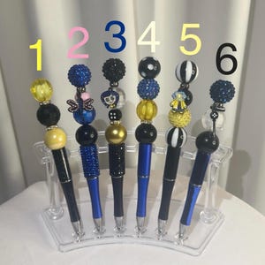May include: Six decorative pens with beaded tops in various colours and designs. The pens have black or blue barrels and are displayed in a clear acrylic stand. The beaded tops include yellow, blue, black, and white accents.
