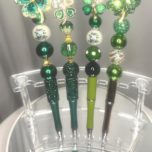 May include: Four decorative pens with shamrock-shaped toppers. The pens feature green and gold accents, with the text "A wee bit Irish" on some. The pens are displayed in a clear acrylic holder.
