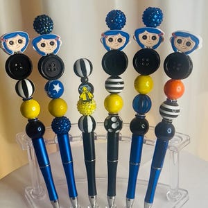 May include: Six decorative pens with unique designs. Each pen has a blue or black barrel and is adorned with beads, buttons, and character toppers. The pens are displayed in a clear acrylic holder. The toppers include a cartoon character.