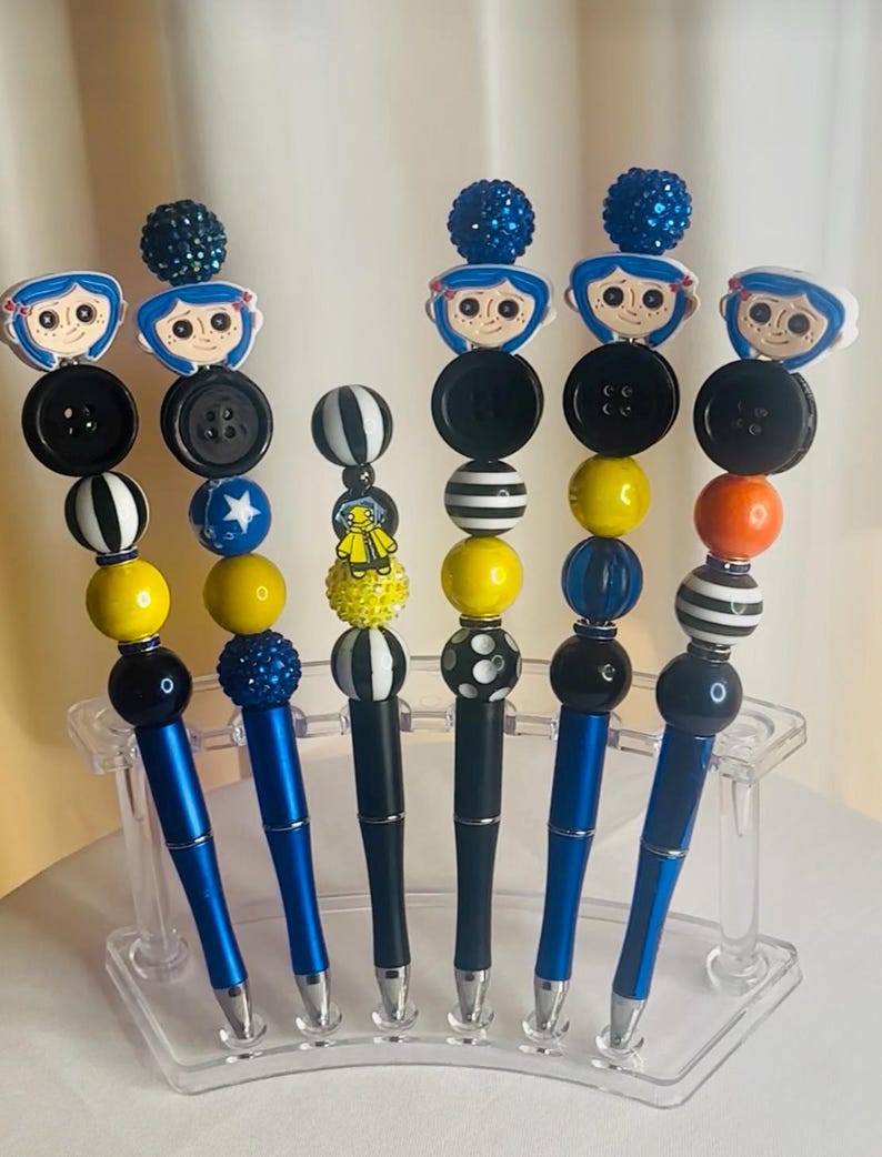May include: Six decorative pens with unique designs. Each pen has a blue or black barrel and is adorned with beads, buttons, and character toppers. The pens are displayed in a clear acrylic holder. The toppers include a cartoon character.