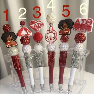 May include: A collection of six decorative pens with various toppers. The pens have red or white barrels, embellished with rhinestones. The toppers include a dress, a heart, a pearl-like sphere, and images of a woman and a sorority logo. The pens are displayed in a clear holder.