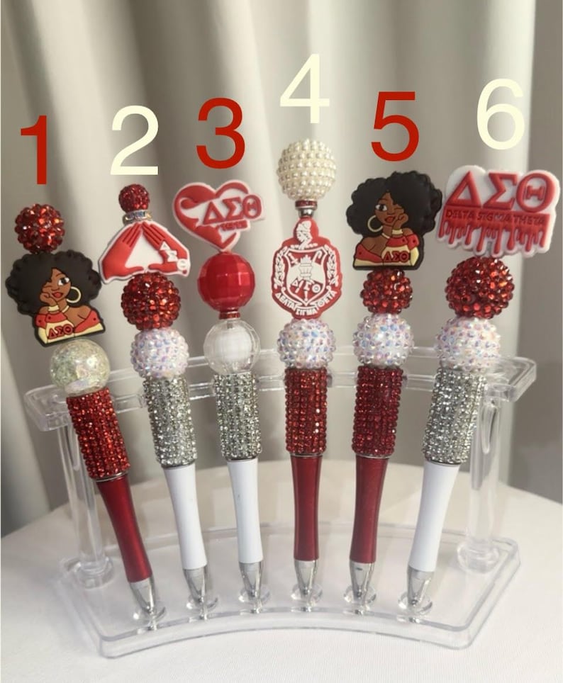 May include: A collection of six decorative pens with various toppers. The pens have red or white barrels, embellished with rhinestones. The toppers include a dress, a heart, a pearl-like sphere, and images of a woman and a sorority logo. The pens are displayed in a clear holder.