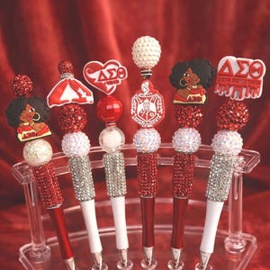 May include: A collection of decorative pens in a clear display case. The pens have red and white barrels, embellished with rhinestones and various toppers, including a heart, a lady, and a crest. The background is a red velvet curtain.