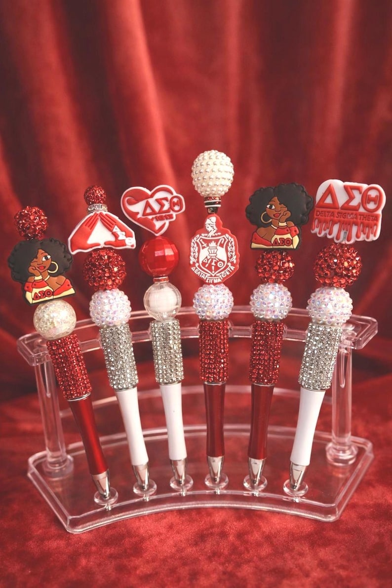 May include: A collection of decorative pens in a clear display case. The pens have red and white barrels, embellished with rhinestones and various toppers, including a heart, a lady, and a crest. The background is a red velvet curtain.