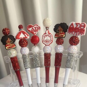 May include: A set of six decorative pens with red and white accents. Each pen features a different charm, including a heart, a dress, and a woman with an afro. The pens are displayed in a clear plastic holder.