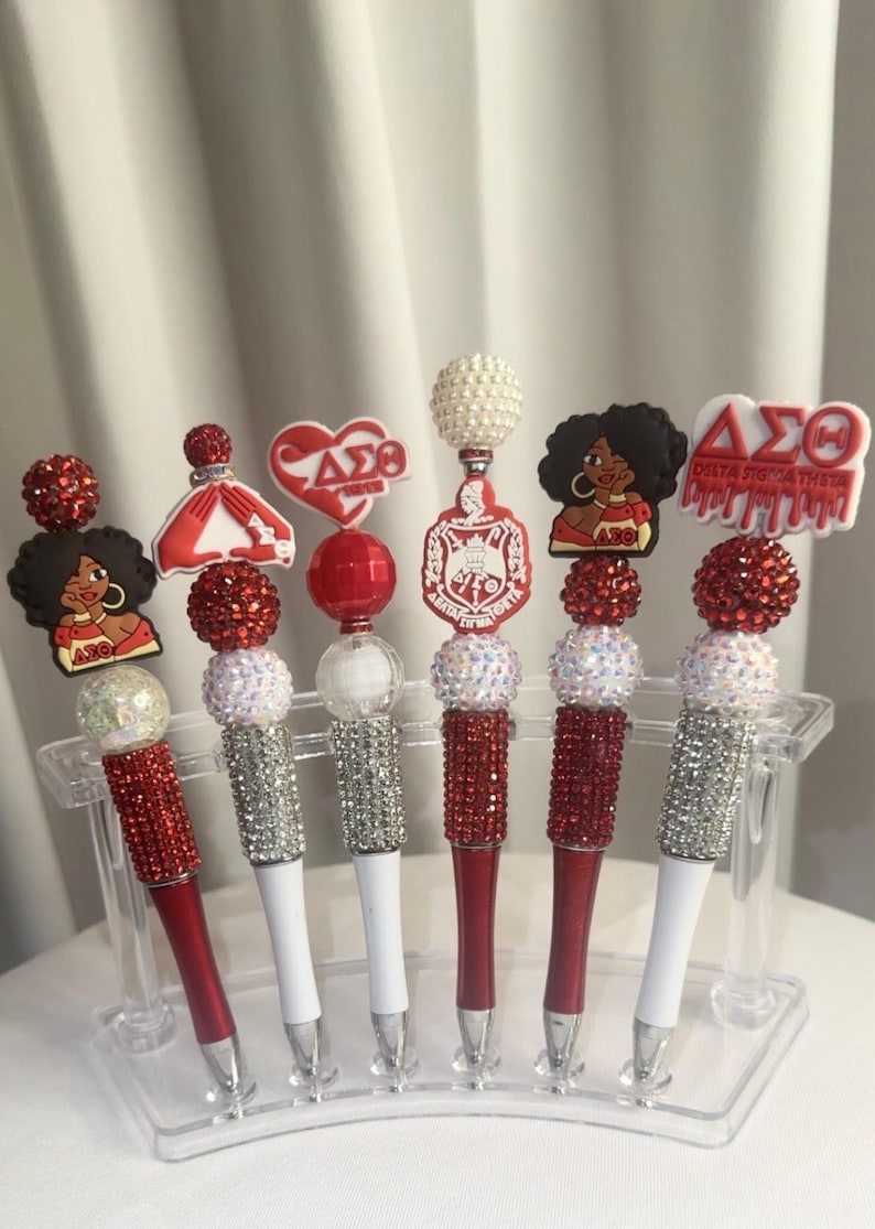 May include: A set of six decorative pens with red and white accents. Each pen features a different charm, including a heart, a dress, and a woman with an afro. The pens are displayed in a clear plastic holder.