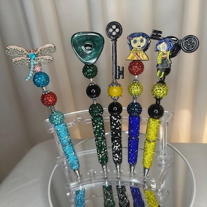 Op de afbeelding: Een verzameling decoratieve pennen met diverse opzetstukken, waaronder een libelle, een groene edelsteen, een sleutel en stripfiguren. De pennen zijn versierd met kleurrijke strass-steentjes en kralen. Ze worden gepresenteerd op een heldere, spiegelende standaard.