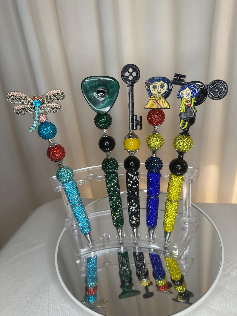 Op de afbeelding: Een verzameling decoratieve pennen met diverse opzetstukken, waaronder een libelle, een groene edelsteen, een sleutel en stripfiguren. De pennen zijn versierd met kleurrijke strass-steentjes en kralen. Ze worden gepresenteerd op een heldere, spiegelende standaard.