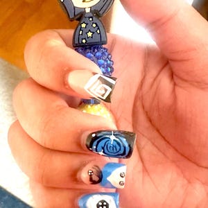 May include: A blue pen with a Coraline-themed topper. The pen is held in a hand with painted nails. The nails feature designs of a button, a spiral, and geometric shapes in blue, black, white, and beige.