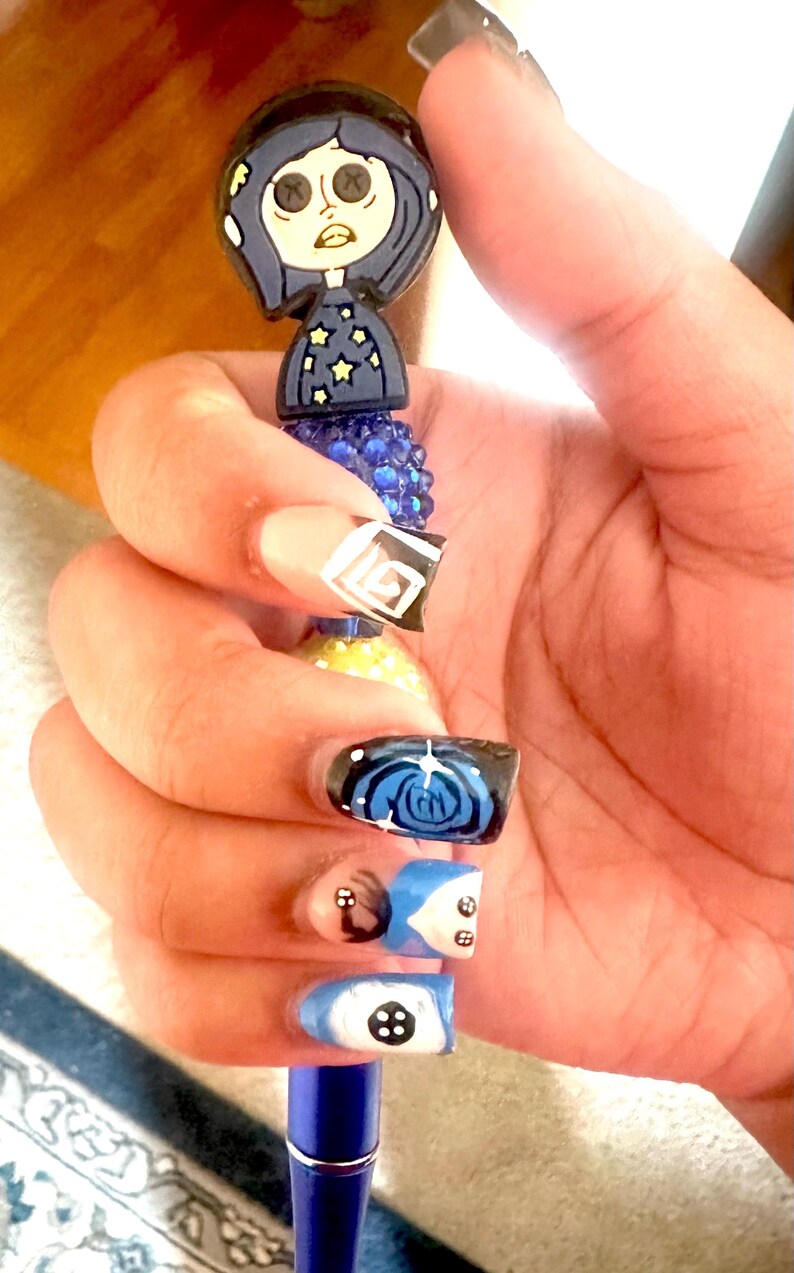 May include: A blue pen with a Coraline-themed topper. The pen is held in a hand with painted nails. The nails feature designs of a button, a spiral, and geometric shapes in blue, black, white, and beige.