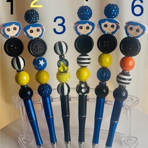 May include: Six decorative pens with blue barrels and various bead and button designs. Each pen features a different topper, including character figures and patterned beads. The pens are displayed in a clear acrylic holder.