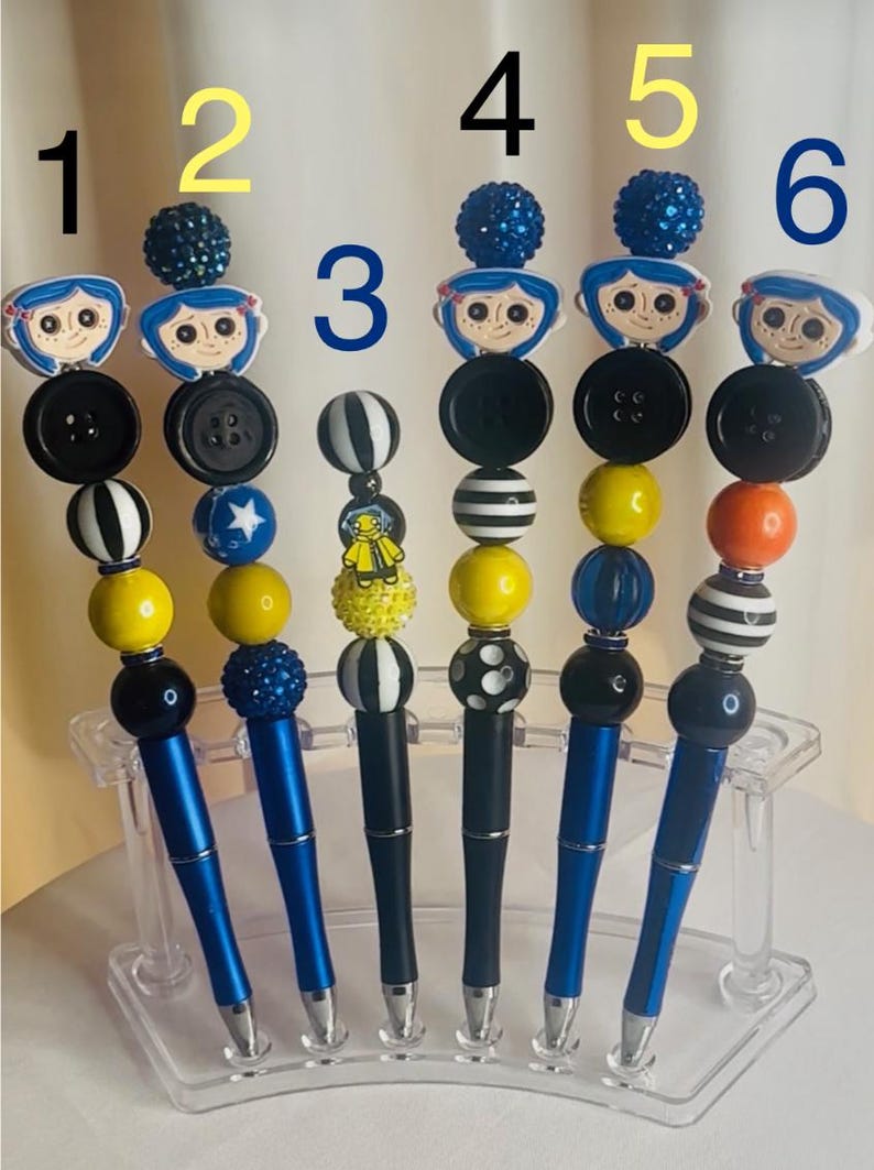 May include: Six decorative pens with blue barrels and various bead and button designs. Each pen features a different topper, including character figures and patterned beads. The pens are displayed in a clear acrylic holder.