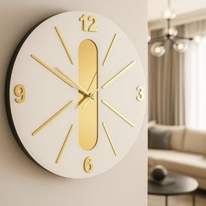 May include: A modern wall clock with a white face and gold-colored accents. The clock features gold hands, hour markers, and the numbers 12, 3, 6, and 9. The clock's face has a gold-colored vertical stripe down the center. The clock is mounted on a light-colored wall.