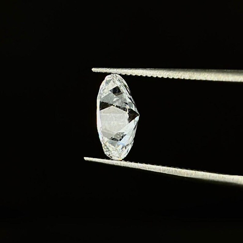 May include: A faceted, oval-shaped diamond held by tweezers against a black background. The diamond is clear and reflects light, showcasing its many facets. The tweezers are silver-colored.