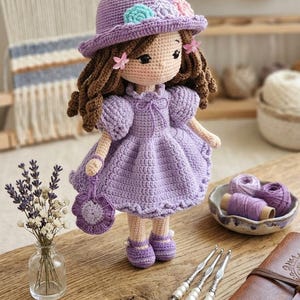 Könnte beinhalten: Handgefertigte Häkelpuppe in einem lavendelfarbenen Kleid und Hut, verziert mit floralen Akzenten. Die Puppe hat braune Locken und eine passende Handtasche. Zubehör umfasst Häkelnadeln, Blumenanhänger und eine kleine Vase mit Lavendel.