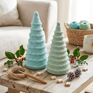 Könnte beinhalten: Zwei handgehäkelte Weihnachtsbäume in Hellblau und Mintgrün stehen auf einem Holztisch. Ein Knäuel Garn, ein Häkelhaken, Holzsterne, Stechpalmen und Tannenzapfen vervollständigen die festliche Szene. Ein Korb mit Garn und eine Tasse sind im Hintergrund.