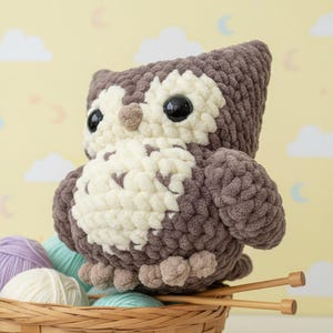 Könnte beinhalten: Ein handgefertigtes Eulen-Plüschtier in Braun- und Cremetönen. Die Eule hat große, schwarze Augen und sitzt in einem Korb mit Garn und Stricknadeln. Der Körper der Eule besteht aus weichem, strukturiertem Garn.