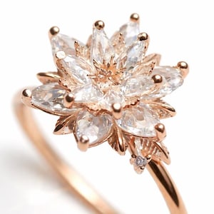 Può includere: Anello in oro rosa con design floreale. Il fiore è composto da pietre chiare taglio marquise, con piccoli accenti dorati sulle punte dei petali. Una piccola pietra rotonda è incastonata vicino alla base del fiore.