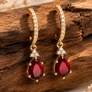 May include: A pair of gold hoop earrings featuring a row of small clear stones along the arch, with a dangling teardrop-shaped red gemstone pendant accented by three tiny clear stones at the top of each drop, creating a refined jewelry piece.