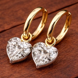 May include: A pair of gold hoop earrings with heart-shaped diamond pendants. The earrings are set against a dark wood grain background, highlighting the sparkle of the diamonds and the shine of the gold.