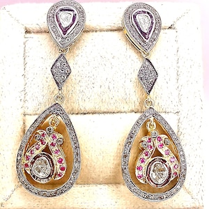 Könnte beinhalten: Ein Paar goldfarbene Ohrhänger mit aufwendigen Details. Jeder Ohrring hat ein tropfenförmiges Design mit einem zentralen Diamanten und roten Edelsteinen. Die Ohrringe haben einen diamantbesetzten Rand und einen rautenförmigen Akzent.