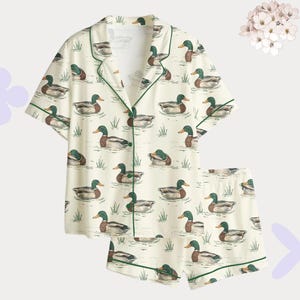 May include: Cream-colored pajama set featuring a repeating pattern of ducks swimming in a pond. The short-sleeved top and shorts are trimmed with green piping. The ducks are depicted in shades of brown, green, and white.