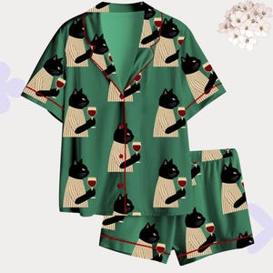 Könnte beinhalten: Grünes Pyjama-Set mit einem sich wiederholenden Muster aus schwarzen Katzen, die Weingläser halten. Das kurzärmlige Oberteil und die Shorts haben rote Paspeln. Die Katzen tragen gestreifte Pullover. Ein lustiges und skurriles Nachtwäsche-Set.