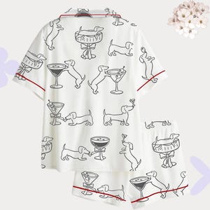 Puede incluir: Conjunto de pijama blanco con un patr&oacute;n de dibujo lineal negro repetido de perros salchicha y copas de martini. El conjunto incluye una camisa de manga corta con botones y pantalones cortos a juego, ambos ribeteados con ribete rojo.