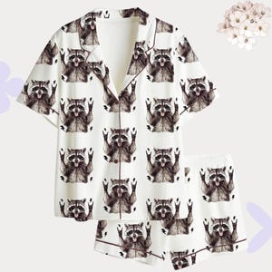 Può includere: Pigiama bianco con un motivo ripetuto di procioni. La maglia a maniche corte e i pantaloncini hanno bordi marroni. I procioni sono raffigurati con le braccia alzate e la lingua di fuori.
