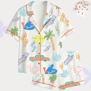 May include: White pajama set with a colourful vacation-themed print. The short-sleeved top and shorts feature illustrations of cars, flamingos, palm trees, and lounge chairs. The top has the text "Vacation" and "Palm Springs".