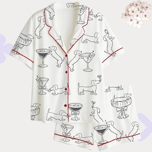 Puede incluir: Conjunto de pijama blanco con un patr&oacute;n repetido de dibujo lineal negro de perros salchicha y copas de martini. El conjunto incluye una camisa de manga corta con botones y pantalones cortos, ambos con ribete rojo.