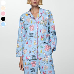 May include: Light blue pajama set with a repeating pattern of Easter-themed nurse-related graphics. The set includes a button-down shirt and pants, trimmed with pink piping. Text includes "Bunny's Nurse" and "#Nurselife".