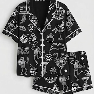 Op de afbeelding: Een zwarte pyjamaset met een shirt met korte mouwen en bijpassende shorts, voorzien van een witte grafische print van skeletten, geesten, pompoenen, discoballen en cocktails. Het shirt bevat de tekst She said yes, witches en LAST BOO.