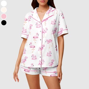 May include: White pyjama set featuring a pink flamingo pattern. The short-sleeved top and shorts are trimmed with pink piping. The button-down top has a notched collar.