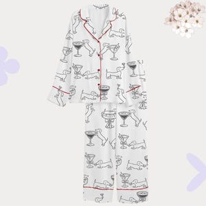 Puede incluir: Conjunto de pijama blanco con un estampado repetido en blanco y negro de perros salchicha y copas de martini. El conjunto incluye una camisa abotonada con ribete rojo y pantalones a juego. Un arreglo floral est&aacute; en el fondo.