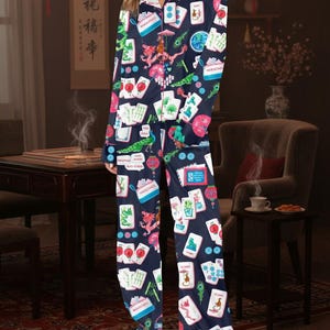 May include: Navy blue pajama set with a colorful print of Mahjong tiles, dragons, and peacocks. The long-sleeved top has a button-down front and a pink floral collar. The matching pants have a relaxed fit. The set is made of a soft, comfortable material.
