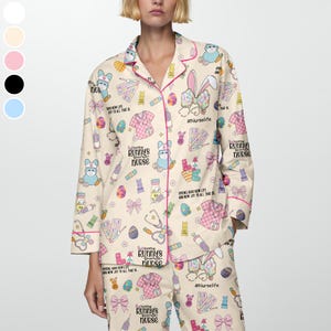 May include: Beige pajama set with a repeating pattern of Easter-themed medical illustrations, bunnies, and text. The set includes a button-down top with pink trim and matching pants. Text includes "Every Bunny's Favorite Nurse" and "#NurseLife."