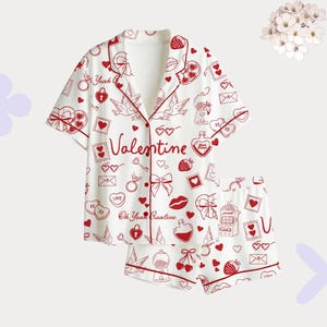 Op de afbeelding: Witte Valentijn pyjamaset met rode bies en een herhaald patroon van harten, strikken en liefdesthema graphics. Het shirt heeft korte mouwen en het woord "Valentine" is erop gedrukt.