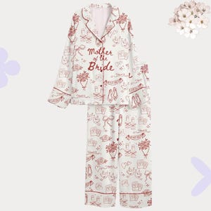 Puede incluir: Un conjunto de pijama blanco de manga larga con botones, ribetes rojos y un patrón repetitivo de líneas rojas con motivos de boda como ramos, palomas, copas de champán y zapatos. El texto Mother of the Bride aparece en el pecho.