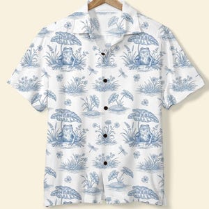 Puede incluir: Camisa blanca con botones con un estampado azul de ranas y botánico. La camisa de manga corta presenta un diseño repetitivo de ranas, plantas y libélulas. La palabra "DAVID" está impresa en las hojas. La camisa está colgada en una percha de madera.